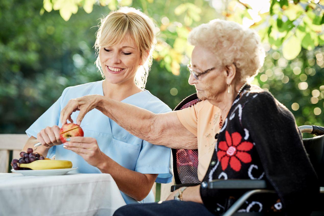 Pflegedienst Guetersloh: Pflegekraft reicht Seniorin im Garten frisches Obst als Teil der taeglichen Betreuung