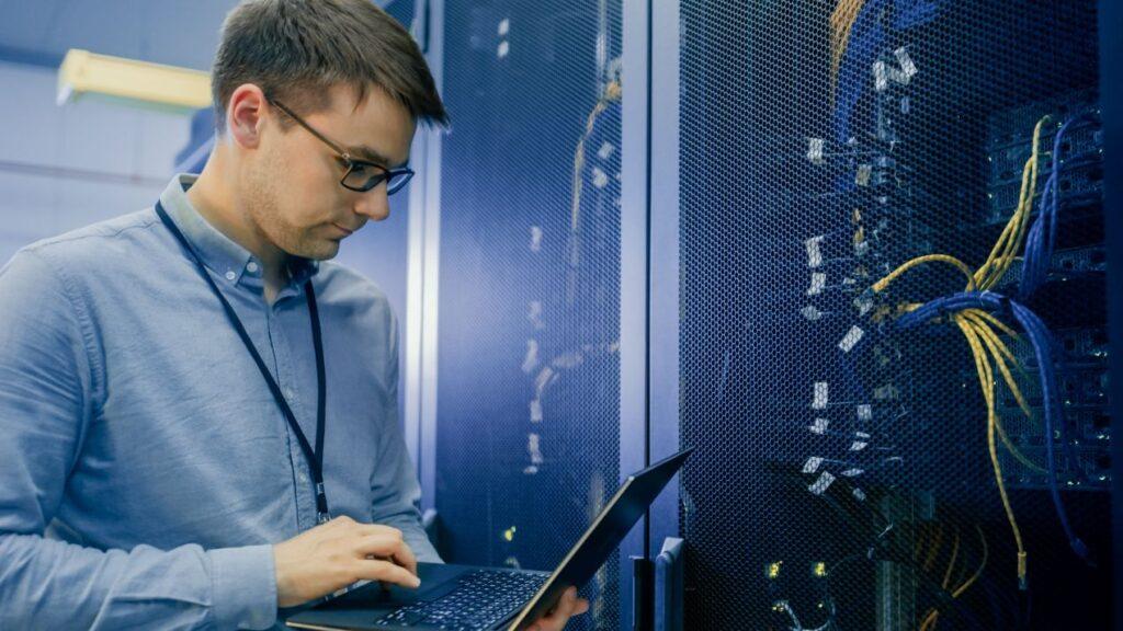 Ein IT-Techniker arbeitet mit Laptop vor einem Serverrack mit MTP-Glasfaserkabeln – Beispiel für moderne Netzwerkinfrastruktur
